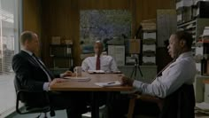 two men sitting at a table talking to each other in an office with papers on the desk