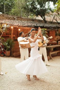 a woman in a white dress dancing with other people around her and one man playing the guitar