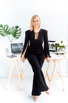 a woman sitting at a desk in front of a laptop computer wearing a black jumpsuit