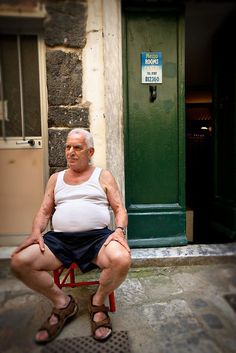 an older man sitting on a red chair in front of a green door and building