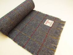 "HARRIS TWEED FRINGED SCARF Handmade in Scotland by The Little Stitch Company  25cm wide 200cm long Keep warm this winter with a cosy Harris Tweed scarf.   At 2 metres long, the scarf is longer than most Harris Tweed scarves.  This gives you greater flexibility in how you wear it.  Keep it casual with one end thrown over your shoulder.  Wrap it round your neck to keep warm and cosy.  Or double it over and pull the ends through the loop for a more modern look.   The scarf is made from handwoven Harris Tweed - a pure, new wool fabric - in a muted blue with a fine overcheck of red and yellow..  It's fringed on all sides with deeper fringing at the ends. It carries the Harris Tweed Authority's orb label which is your guarantee of authenticity - please scroll down for more information about Har