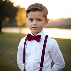 a young boy wearing a bow tie and suspenders standing in front of a lake