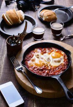 an iron skillet filled with eggs on top of a wooden table next to bread