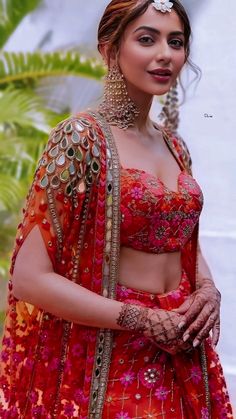 a woman in a red and orange outfit with jewelry on her head, standing next to a palm tree