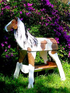 a toy horse sitting on top of a wooden bench in the grass next to purple flowers