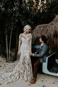 a man standing next to a woman in a wedding dress near a car with thatched roof