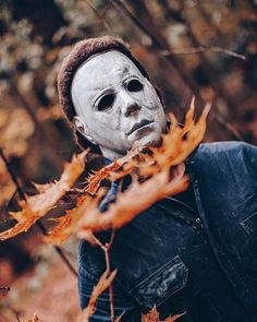 a man wearing a white mask standing in the woods with autumn leaves around his neck