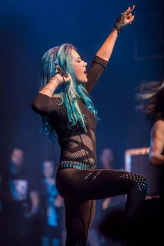 lady with blue hair performing on stage in black outfit and leggings holding up her hand