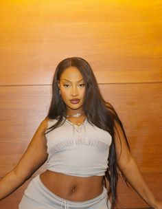 a woman with long hair standing in front of a wooden wall wearing a white top