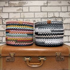 two knitted baskets sitting on top of a brown suitcase next to a brick wall