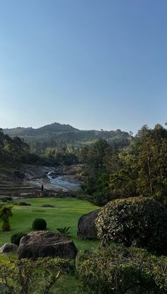 a lush green field surrounded by trees and rocks with a river running through it in the distance