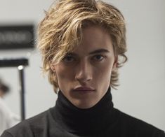 a young man with blonde hair wearing a black turtle neck sweater looking at the camera