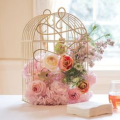 a birdcage filled with pink flowers next to a wine glass and napkins