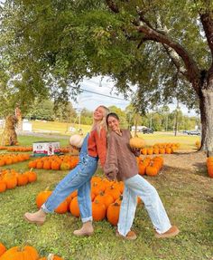 two women are posing in front of pumpkins on the ground with trees behind them