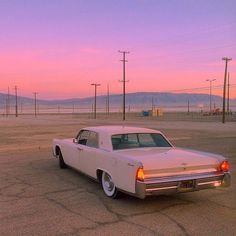 an old white car parked in a parking lot with the sun setting on it's horizon