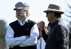 two men standing next to each other with their arms crossed and one man wearing a hat