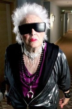 an older woman with white hair and sunglasses on her face wearing a black jacket, purple top and silver pants