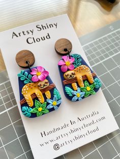 the earrings are decorated with colorful flowers and slotty bears on wooden hooks, sitting on a card that says artsy shiny cloud