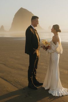 a bride and groom standing on the beach looking at each other