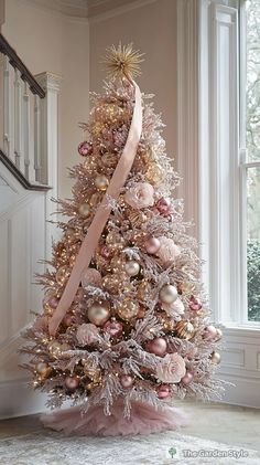 a decorated christmas tree with pink and gold ornaments in front of a window, next to a staircase