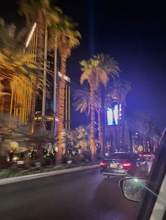 cars driving down the street at night with palm trees and tall buildings in the background