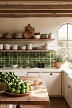 a wooden table topped with lots of green fruit next to a kitchen counter covered in white dishes