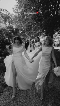 two women in white dresses are holding hands and walking down the street with other people behind them