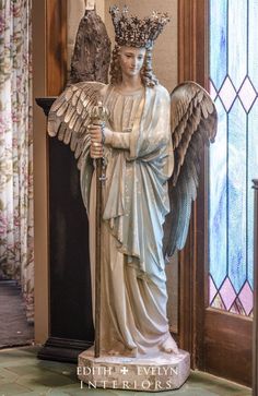 a statue of an angel holding a cross in front of a stained glass window with floral curtains