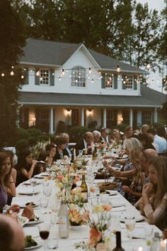 a group of people sitting at a long table with food and drinks in front of a house