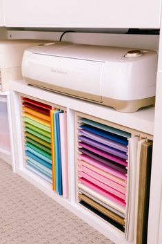 a printer sitting on top of a white shelf filled with colored papers and folders