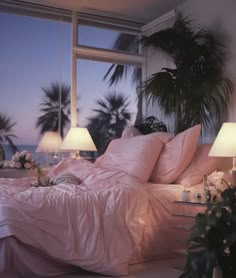 a bed with pink sheets and pillows in front of a window at night, next to potted palm trees