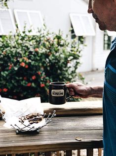 a man holding a coffee cup on top of a wooden table next to a plate of food