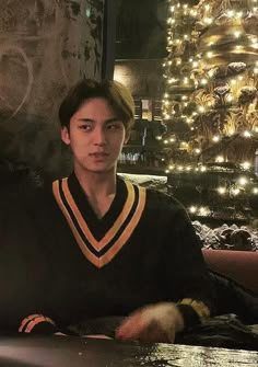 a young man sitting at a table in front of a christmas tree with lights on it