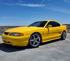 a yellow mustang parked in a parking lot next to a wall and blue sky with wispy clouds