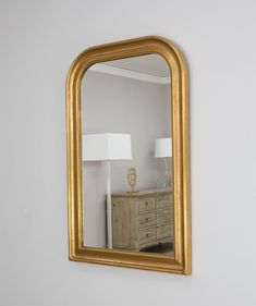 a gold framed mirror hanging on the wall above a dresser with a lamp next to it