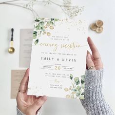 two hands holding up a wedding reception card with gold foil lettering and greenery on it