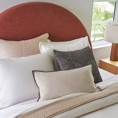 a bed topped with lots of pillows next to a night stand and window sill