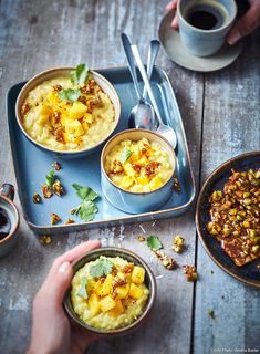 three bowls of food are on a tray with spoons and two cups of coffee