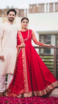 a man and woman standing next to each other on a balcony wearing red dresses with gold accents