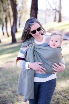 a woman holding a baby wrapped in a grey and white striped wrap with trees in the background