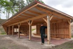 a man standing in front of a wooden structure on top of a dirt field next to trees