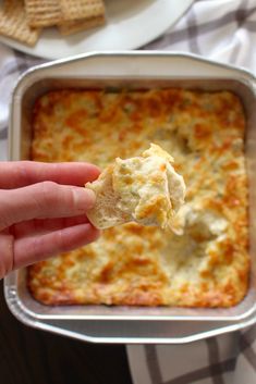 a hand holding a cracker over a casserole dish with cheese on it