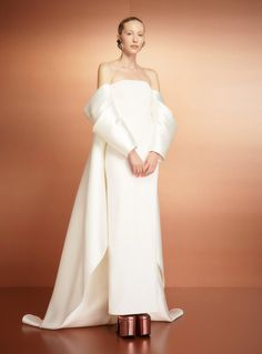 a woman standing in front of a brown background wearing a white dress with an asymmetrical neckline