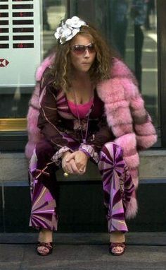 a woman sitting on top of a bench in front of a building wearing sunglasses and a fur coat