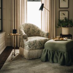 a living room with a chair, ottoman and table in front of a window filled with curtains