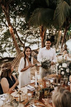 a group of people standing around a table with food and drinks on it, surrounded by palm trees