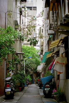 an alley way with parked scooters and clothes hanging on the buildings, trees and plants