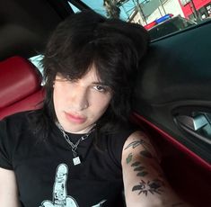 a man with black hair and tattoos sitting in a car looking at the camera while holding his hand up