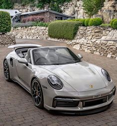 a silver porsche sports car parked in front of a stone wall and shrubbery behind it