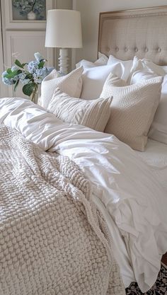 a white bed topped with lots of pillows and blankets next to a lamp on a table